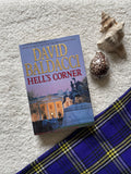 Book titled 'Hell's Corner' by David Baldacci on a textured surface with shells and a plaid fabric.