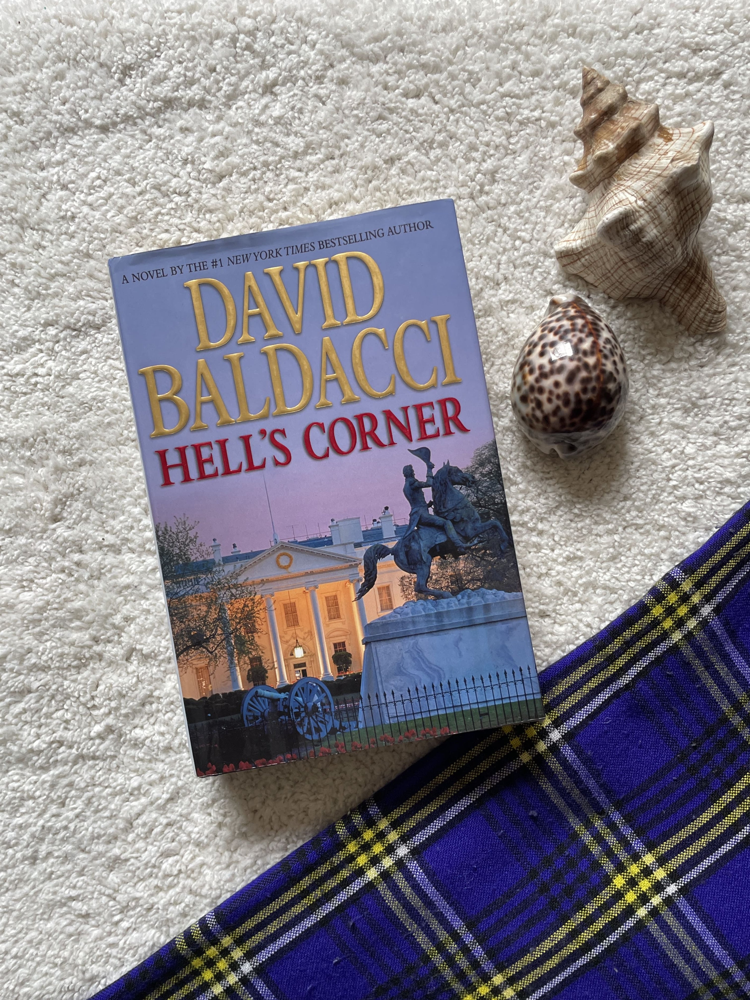 Book titled 'Hell's Corner' by David Baldacci on a textured surface with shells and a plaid fabric.