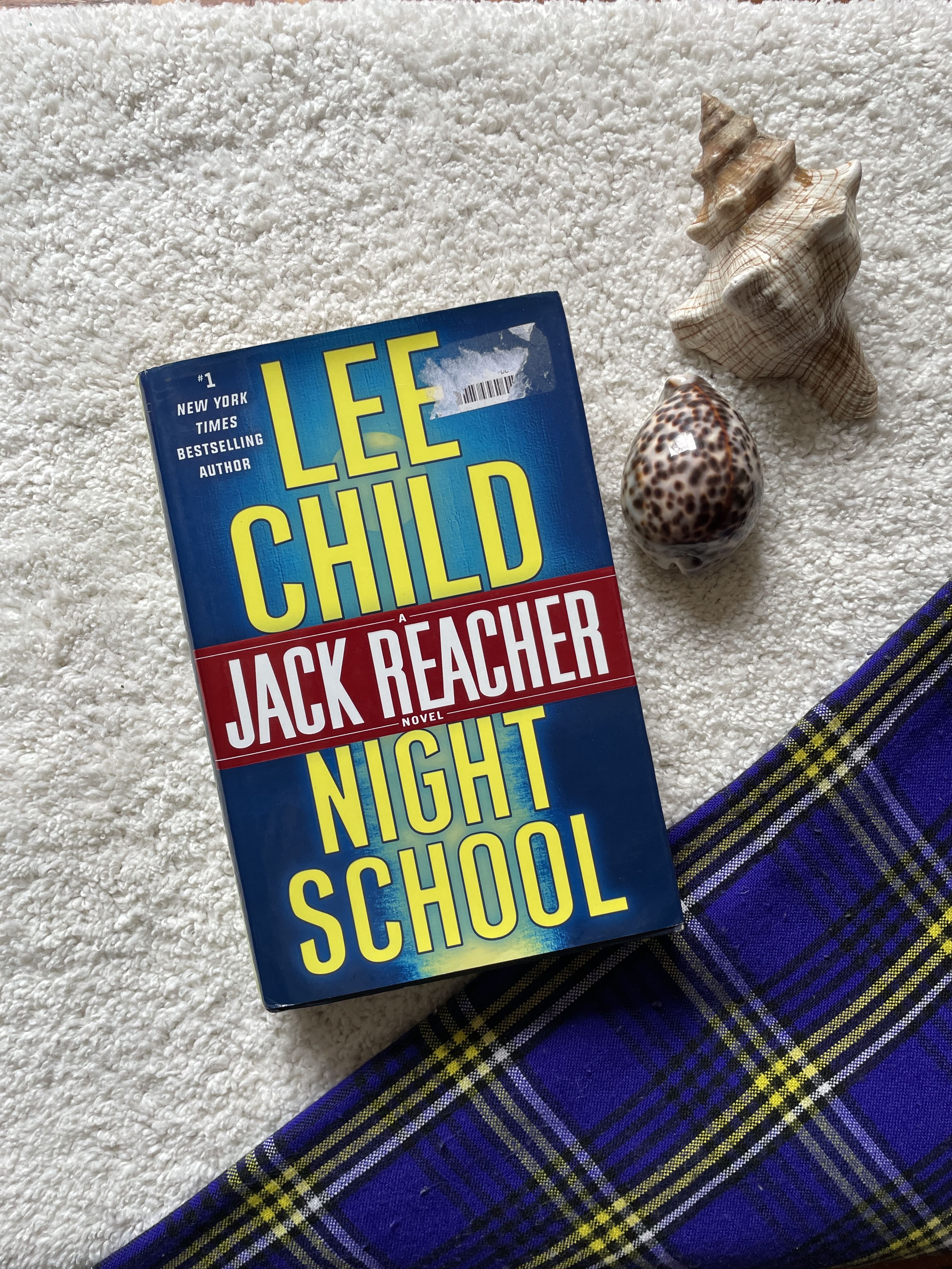 Book titled 'Night School' by Lee Child and Jack Reacher on a textured surface with shells and fabric.