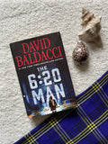 Book titled 'The 6:20 Man' by David Baldacci on a textured surface with shells and fabric.