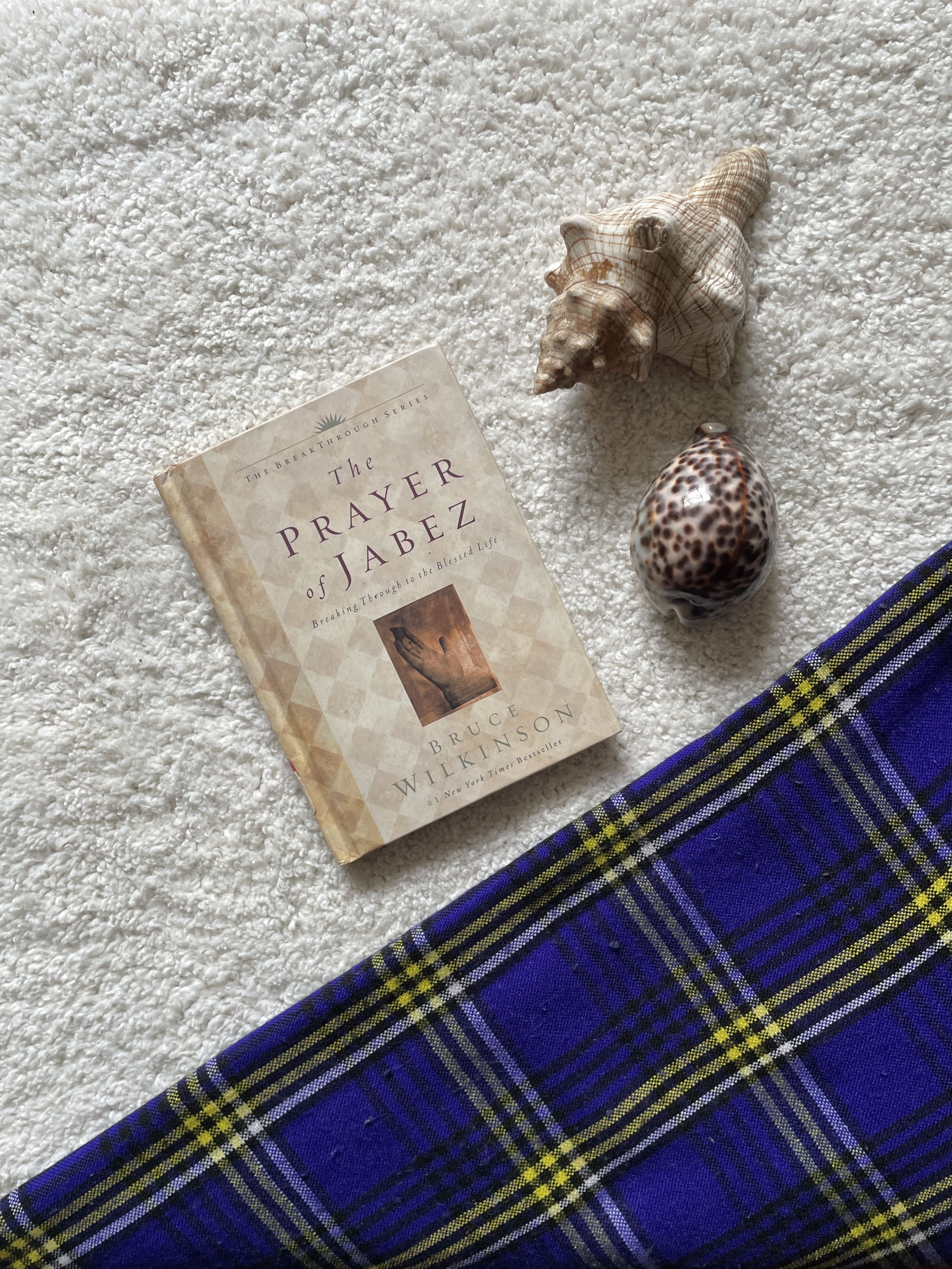 Book titled 'The Prayer' with seashells on a textured surface next to a blue and yellow checkered fabric.